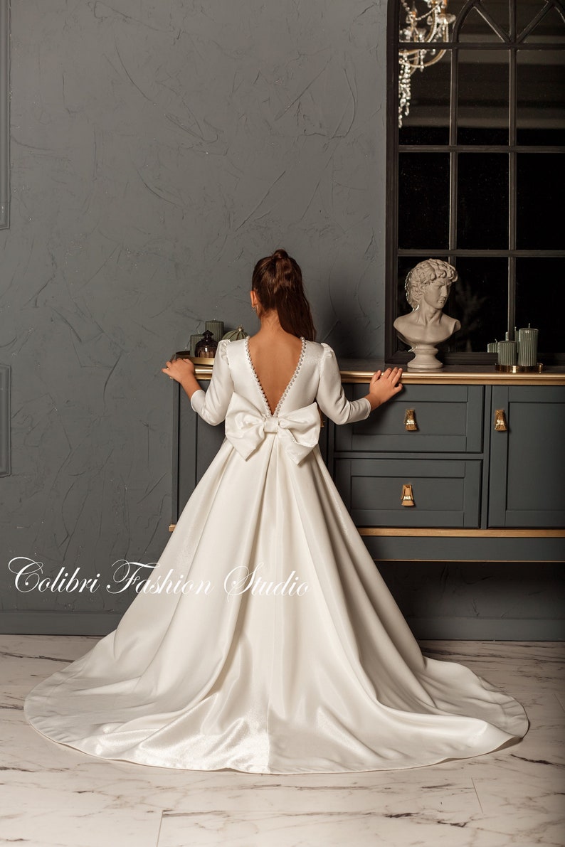 Classic ivory dress for a flower girl. The dress is made of satin. The top is decorated with pearls along the neckline and has satin sleeves. The skirt is satin and floor length. The back of the dress is complemented by a satin bow.
