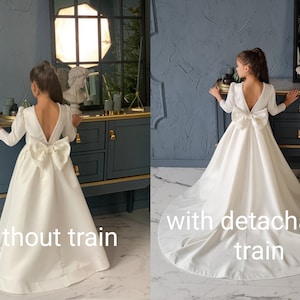 Classic ivory dress for a flower girl. The dress is made of satin. The top is decorated with pearls along the neckline and has satin sleeves. The skirt is satin and floor length. The back of the dress is complemented by a satin bow.