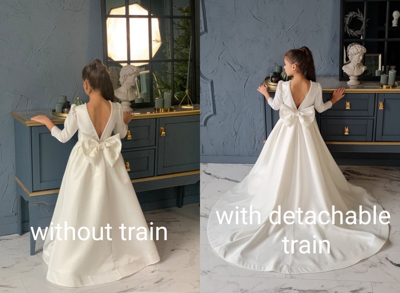 Classic ivory dress for a flower girl. The dress is made of satin. The top is decorated with pearls along the neckline and has satin sleeves. The skirt is satin and floor length. The back of the dress is complemented by a satin bow.