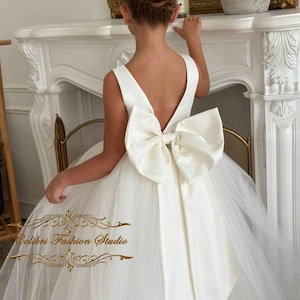 May include: A white tulle and satin dress with a large bow on the back. The dress has a V-shaped back and is sleeveless. The dress is for a young girl.