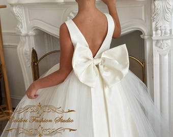 May include: A white tulle and satin dress with a large bow on the back. The dress has a V-shaped back and is sleeveless. The dress is for a young girl.