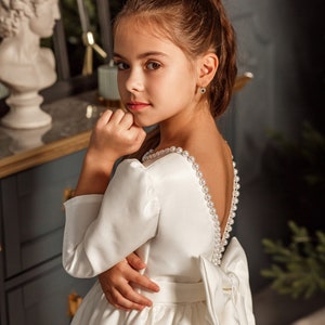 Classic ivory dress for a flower girl. The dress is made of satin. The top is decorated with pearls along the neckline and has satin sleeves. The skirt is satin and floor length. The back of the dress is complemented by a satin bow.