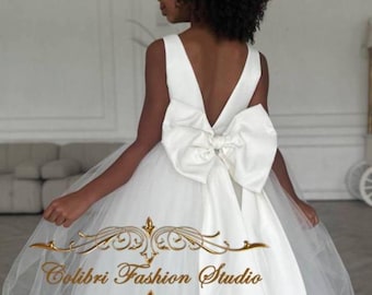 May include: A young girl wearing a white tulle dress with a large white satin bow on the back. The dress has a V-shaped neckline and sleeveless straps. The girl has curly black hair and is standing in a room with white walls. The text "Colibri Fashion Studio" is visible on the dress.