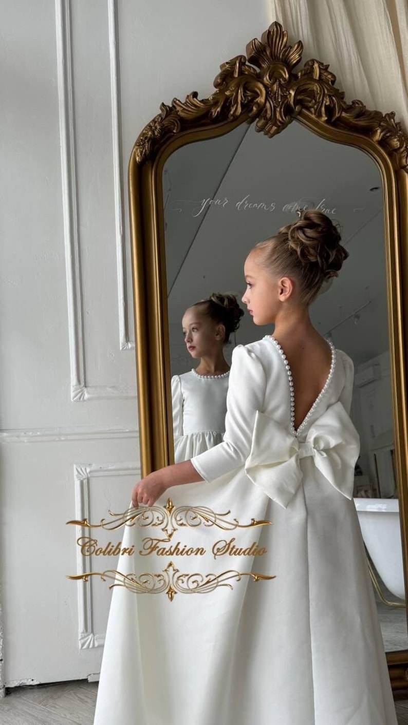 Classic ivory dress for a flower girl. The dress is made of satin. The top is decorated with pearls along the neckline and has satin sleeves. The skirt is satin and floor length. The back of the dress is complemented by a satin bow.