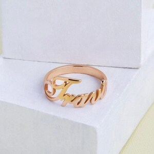 Puede incluir: Un anillo de oro rosa con el nombre "Travis" escrito en cursiva.