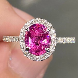 Puede incluir: Un anillo de plata con una gran gema rosa ovalada, rodeada por un halo de gemas claras más pequeñas. La banda del anillo también está adornada con pequeñas gemas claras, creando un efecto brillante. El anillo está sobre un fondo neutro.