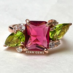 Op de afbeelding: Een ring van roségoud met een grote vierkante roze edelsteen, omringd door kleinere heldere en groene edelstenen. De ring is bezet met een peervormige groene edelsteen aan elke kant van de roze edelsteen. De ring is gelabeld met "AYAN GEMS".