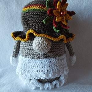 Puede incluir: Muñeca de ganchillo hecha a mano con un sombrero marrón adornado con una flor colorida y un borde amarillo. La muñeca tiene una falda blanca con un borde con volantes y una nariz beige. Los brazos y pies de la muñeca son grises con puntas blancas.