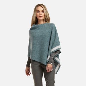 Könnte beinhalten: Ein tealblauer Poncho mit weißen Streifen an den Rändern. Der Poncho wird über einem langärmeligen Shirt und einer grauen Hose getragen.