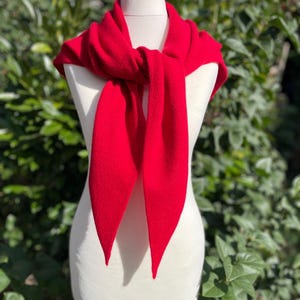 May include: A bright red scarf with a smooth, solid-coloured knit texture. The scarf is tied in a simple knot at the centre, featuring two long, pointed ends that drape downward. The fabric appears soft and uniform in colour throughout the entire piece.