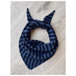 May include: A blue and black striped scarf, knotted at the top, rests on a wooden surface. The scarf is triangular, with horizontal stripes. The ends are pointed, creating a distinctive design. The scarf is made of soft material.