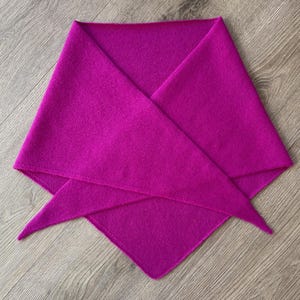 May include: A bright magenta scarf, folded into a triangular shape. The scarf is made of a soft, textured fabric and is displayed on a light-coloured wooden surface. The scarf's edges are neatly finished, highlighting its quality.