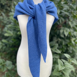 May include: A solid blue knit scarf draped around a white mannequin. The scarf features a soft, textured fabric with a triangular shape that tapers to pointed ends, tied in a simple knot at the centre to create a layered, draped appearance.