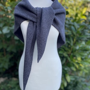 May include: A dark grey triangular shawl with a textured knit finish draped over a white mannequin. The shawl is tied in a simple knot at the centre, with two long pointed ends hanging down, showcasing the soft fabric and consistent charcoal colour.