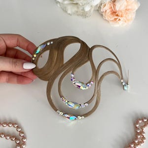 May include: A light brown hair accessory featuring curved, wavy strands of synthetic hair arranged in a swirling pattern. The hair is adorned with iridescent, multi-faceted crystals in various sizes, including marquise and round shapes.