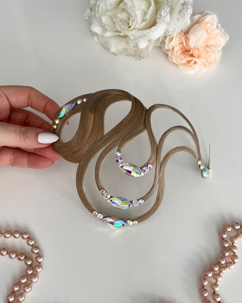May include: A light brown hair accessory featuring curved, wavy strands of synthetic hair arranged in a swirling pattern. The hair is adorned with iridescent, multi-faceted crystals in various sizes, including marquise and round shapes.