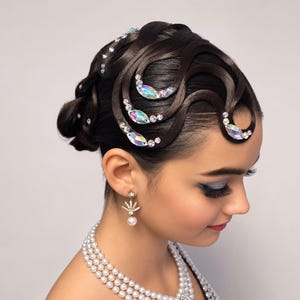 May include: A dark brown hair updo styled in intricate, smooth waves adorned with iridescent, oval-shaped crystals and small round rhinestones. The hairstyle is complemented by gold-toned leaf-shaped earrings with a dangling pearl and a multi-strand pearl necklace.