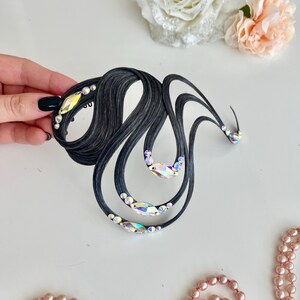 May include: A black hair accessory featuring curved, wave-like strands arranged in an abstract shape. The piece is embellished with multiple iridescent, multi-faceted crystals of varying sizes placed along the dark, textured lines of the design.