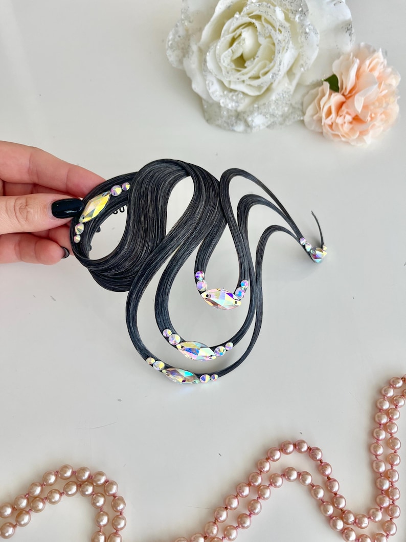 May include: A black hair accessory featuring curved, wave-like strands arranged in an abstract shape. The piece is embellished with multiple iridescent, multi-faceted crystals of varying sizes placed along the dark, textured lines of the design.