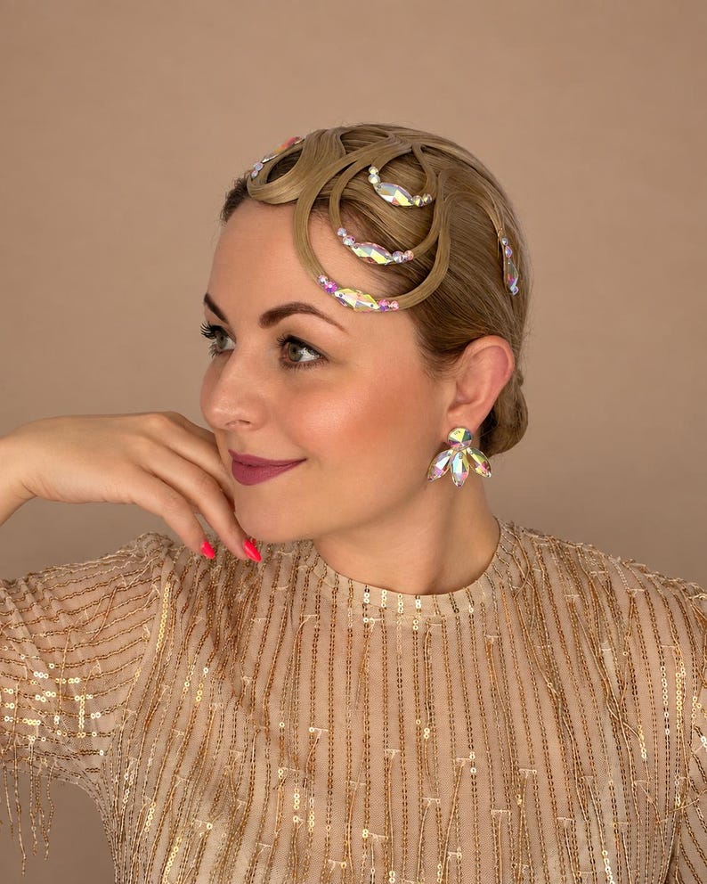 May include: A hair accessory featuring light brown hair styled in intricate loops adorned with iridescent, multi-faceted crystals. The accessory is paired with matching crystal stud earrings that feature a cluster of marquise-cut stones.
