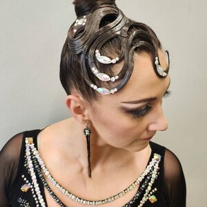 May include: A dark brown hair bun styled with intricate, sculpted waves across the forehead, adorned with iridescent crystals and small white beads. The hair is paired with a black garment featuring a neckline embellished with rows of crystals and beads.