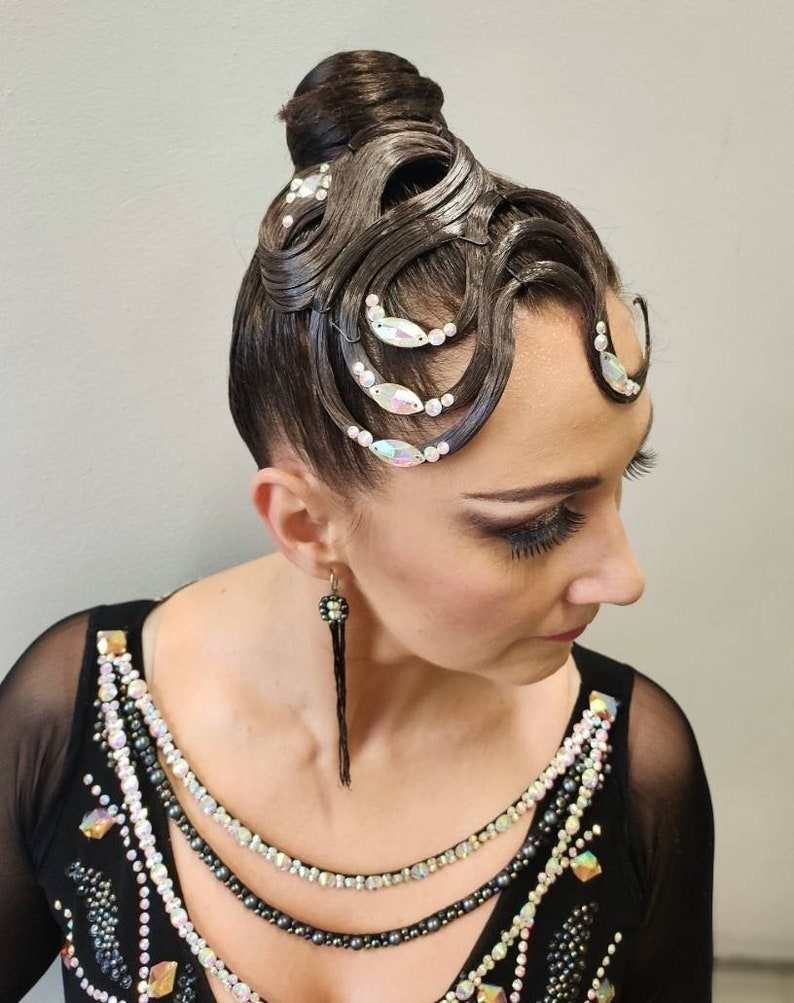 May include: A dark brown hair bun styled with intricate, sculpted waves across the forehead, adorned with iridescent crystals and small white beads. The hair is paired with a black garment featuring a neckline embellished with rows of crystals and beads.