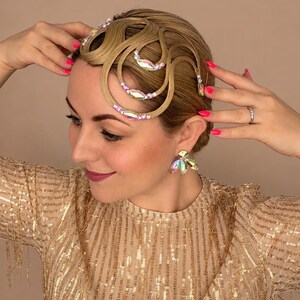 May include: A blonde hair accessory featuring intricate, curved, wave-like hair sections adorned with iridescent, multi-faceted crystals and small white beads. The accessory is paired with matching crystal-embellished earrings in a floral shape.