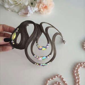 May include: A brown hair accessory featuring a series of curved, looped strands of hair arranged in an intricate, flowing design. The piece is embellished with several iridescent, marquise-shaped crystals and smaller round clear rhinestones.