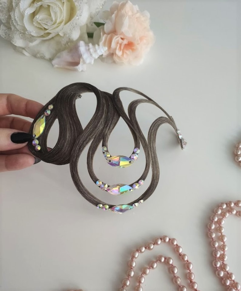 May include: A brown hair accessory featuring a series of curved, looped strands of hair arranged in an intricate, flowing design. The piece is embellished with several iridescent, marquise-shaped crystals and smaller round clear rhinestones.