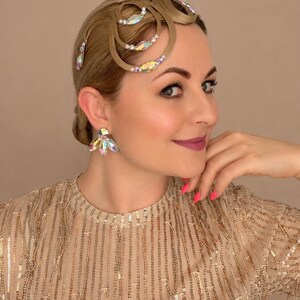May include: A hair accessory featuring curved, light brown hair strands arranged in loops, embellished with iridescent, multi-faceted crystals and small white beads. The accessory is paired with matching iridescent crystal stud earrings.