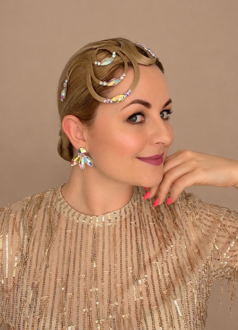 May include: A hair accessory featuring curved, light brown hair strands arranged in loops, embellished with iridescent, multi-faceted crystals and small white beads. The accessory is paired with matching iridescent crystal stud earrings.