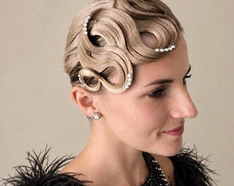 May include: A blonde hairstyle featuring sculpted finger waves arranged in a decorative pattern across the side of the head. The hair is adorned with small, clear, round rhinestones placed along the curves of the waves. A single rhinestone stud earring is visible.
