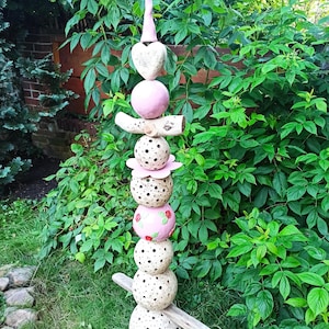 Könnte beinhalten: Eine skurrile Gartenskulptur aus gestapelten Keramikelementen. Sie besteht aus einer rosa konischen Spitze, einem herzförmigen Element und Kugeln mit gepunkteten und floralen Mustern. Holzäste sind integriert, vor dem Hintergrund üppigen Grüns.