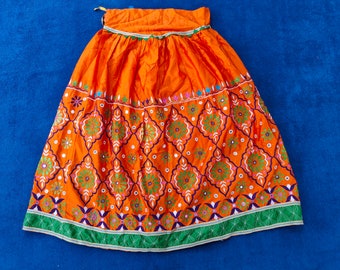 May include: An orange skirt with intricate embroidery and mirror work. The skirt has a green, blue, and white trim around the bottom.
