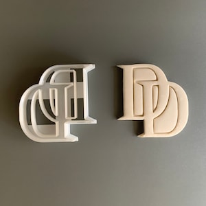May include: Two cookie cutters, one white and one beige, each shaped with the letters "DD" in a stylized font. The white cutter is a negative space design, while the beige one is solid. The cutters are likely for baking.