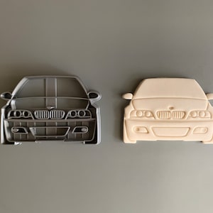 Puede incluir: Un par de cortadores de galletas con forma de un BMW M3 visto de frente. El cortador de la izquierda es gris y el de la derecha es blanco.