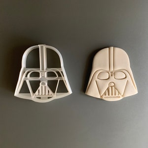 May include: A white plastic cookie cutter and a beige cookie in the shape of Darth Vader's helmet.