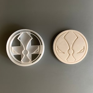 May include: A white plastic cookie cutter in the shape of a stylised logo with a cross and a circle in the centre. The cutter is shown next to a solid white plastic version of the same logo.