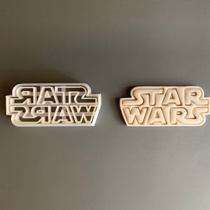 May include: Two white plastic cookie cutters shaped like the Star Wars logo. The top cutter has a striped design, while the bottom cutter is solid.