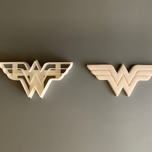 Könnte beinhalten: Zwei weiße Plastik-Ausstecher in Form des Wonder Woman-Logos. Der Ausstecher links ist ein detailliertes Design mit mehreren Schichten. Der Ausstecher rechts ist ein einfacheres Design mit einer einzigen Schicht.