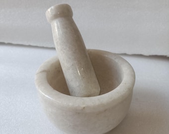 May include: White marble mortar and pestle set. The mortar is a bowl shape with a slightly raised rim. The pestle is a cylinder shape with a rounded end.