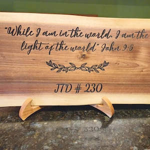 May include: A wooden sign with the quote "While I am in the world. I am the light of the world" from John 9:5, along with a leaf design and "JTD #230". The sign is displayed on a wooden stand.