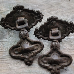 May include: Two antique bronze drawer pulls with ornate, scrollwork designs. Each pull features a decorative backplate, a central handle, and a detailed lower section. The pulls are likely made of cast metal and have a weathered appearance.
