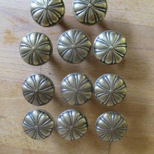 May include: Twelve gold-toned metal cabinet knobs with a decorative floral design. Each knob has a raised centre and a slightly rounded shape.