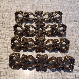 May include: Four ornate gold-toned metal drawer pulls with a scrolled design. The pulls are arranged in a stack, with the top pull facing the viewer.