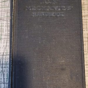 May include: A dark blue, vintage book with the title "I.C.S. Mechanics' Handbook" embossed on the cover. The book's cover has a textured finish and shows signs of age. The background is a patterned surface, suggesting it is an antique or a used item.