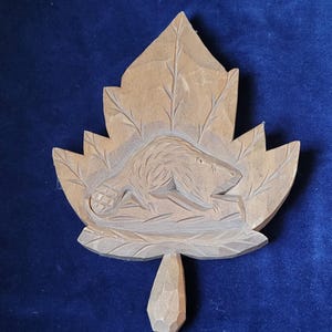 May include: A wooden carving of a beaver on a maple leaf. The beaver is carved in detail, with visible fur and features. The leaf has detailed veins and a textured surface. The carving is a light brown color.
