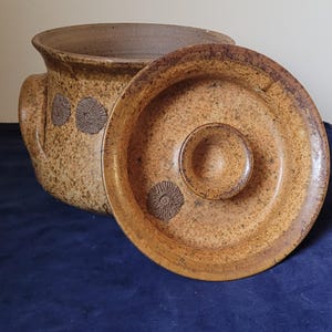 May include: A ceramic casserole dish with a lid. The dish is a speckled tan colour with dark brown floral designs and a handle on each side. The lid is a similar colour with a central knob and a floral design. The dish is open, revealing the interior.