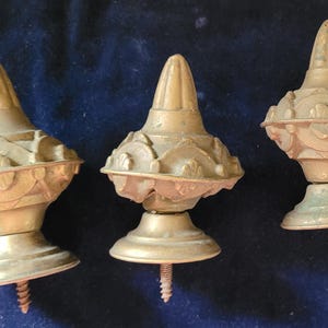 May include: Three antique brass finials with ornate, detailed designs. Each finial has a conical top, a decorative midsection, and a circular base with a screw. The finials are arranged against a dark blue background.