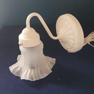 May include: Off-white wall sconce with a frosted glass shade. The shade has a ruffled edge and a floral pattern. The sconce features a curved arm and a decorative backplate. The fixture is designed for wall mounting and has a wire for electrical connection.